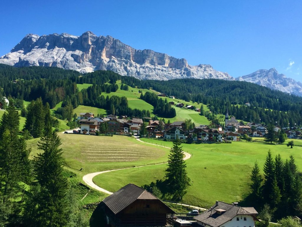 A panoramic view of Badia in Alta Badia, featuring traditional houses, green meadows, and the Dolomites in the background on a sunny day
