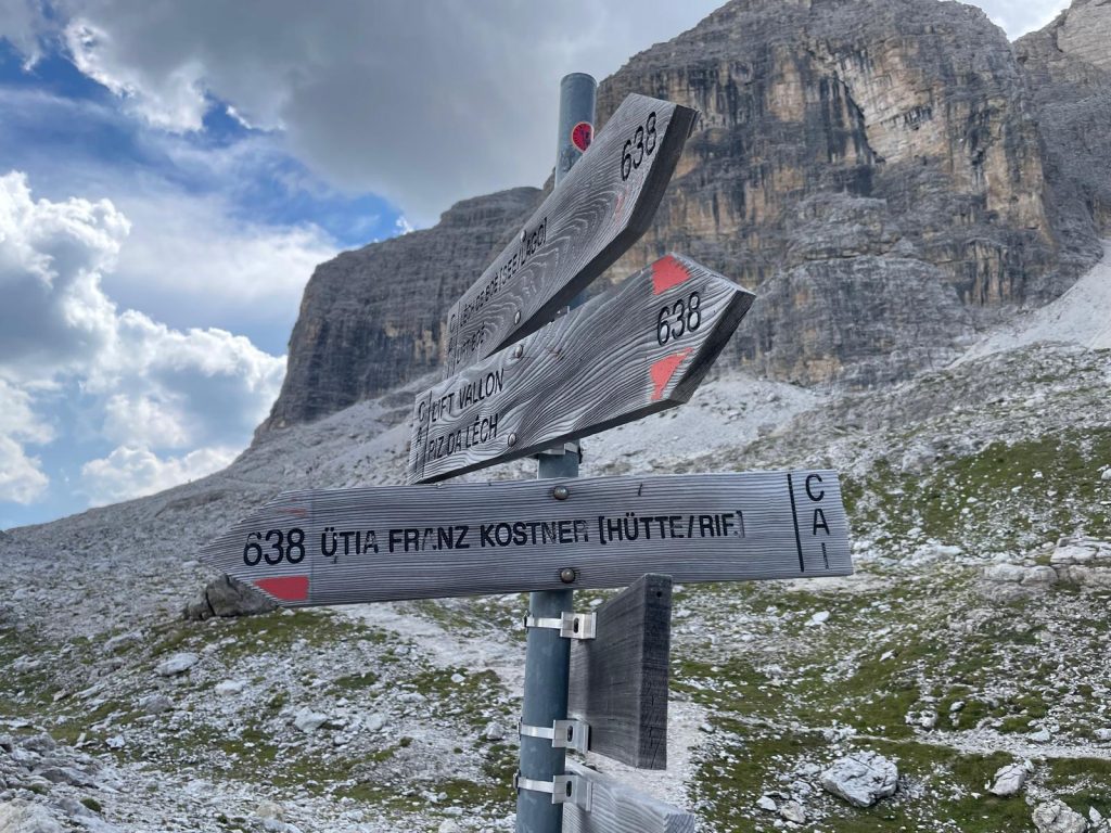 Signpost for Alta Badia via ferrata trails with directions to Ütia Franz Kostner, featuring the Dolomites mountain landscape in the background
